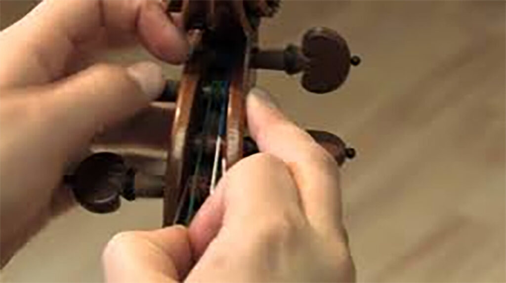 Close-up of hands tuning a violin. One hand pinches the string while the other hand turns a peg on the instrument's scroll. The background is blurred.