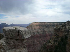 A wide view of the Grand Canyon, showcasing its layered red rock formations and a cloudy sky above. A large, flat rock juts out in the foreground, overlooking the expansive canyon landscape.