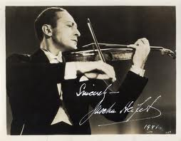 A black and white photograph of a man in a tuxedo playing the violin. His head is tilted slightly, focusing on the strings. The image features a cursive signature at the bottom.