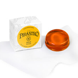 A Pirastro Gold Rosin sits beside its ornately designed white box on a plush cloth. This amber-colored, round rosin is frequently favored by string musicians for enhancing the bowing of their instruments.