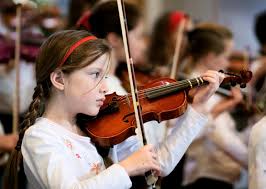 A young girl plays the violin, focusing intently on her music. She wears a white shirt and a red headband. Other children with violins are blurred in the background, indicating a group setting, possibly a practice or performance.