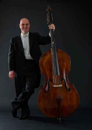 A man wearing formal attire, including a black tuxedo and white bow tie, stands with a double bass in an upright position. He rests his hand on the instrument against a dark background.