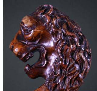 A carved wooden sculpture depicting the profile of a lion's head with detailed mane and open mouth, set against a dark background.