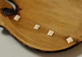 Close-up of a wooden board with four small square pieces of paper or tape attached in a row near the edge. The wood has a natural grain pattern and a slightly worn appearance.