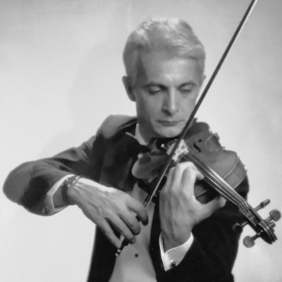 A grayscale image of a man in a suit playing the violin. He is focused on the instrument, with the bow in his right hand and the violin held under his chin. The background is plain and blurred.